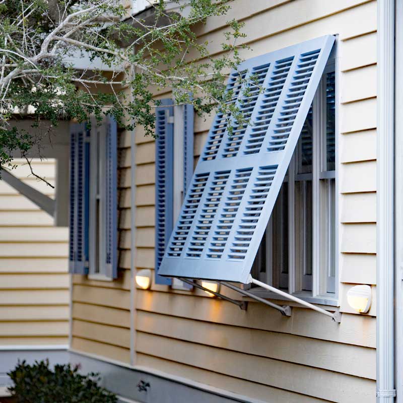 Blue PVC Bahama shutters supported with shutter stays and a top hinge.