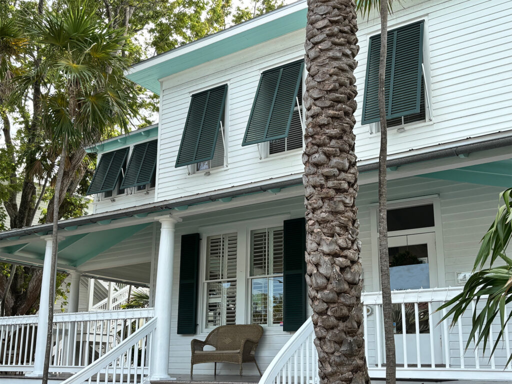 Green aluminum Bahama shutters on the second story of a house with louvered colonial hurricane shutters on the lower level.