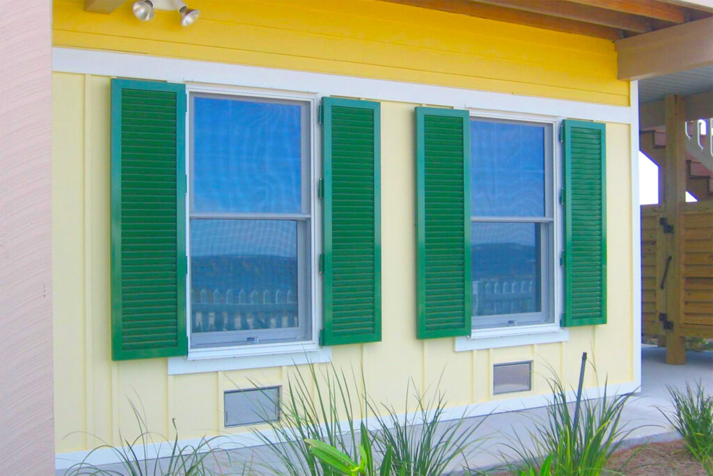 Green aluminum colonial hurricane shutters installed on hinges to a yellow house.